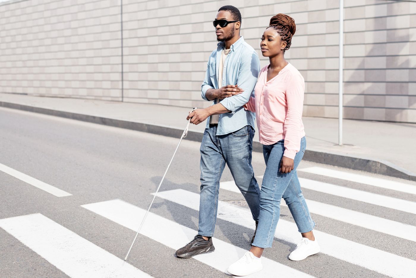 A blind man with a white cane crossing a road arm in arm with a woman